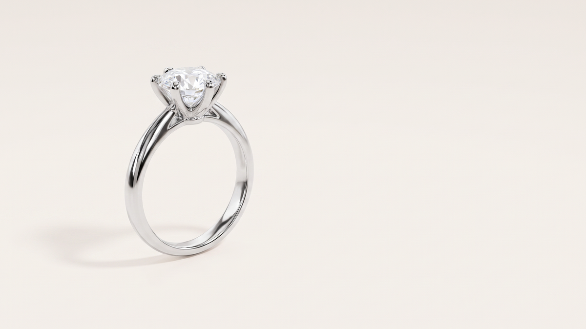 A silver six-prong solitaire ring with a round colourless stone, standing upright on a warm off white background.