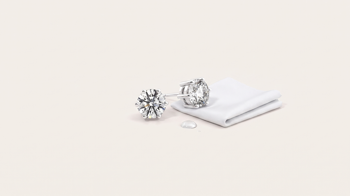 A pair of round six-prong moissanite stud earrings resting beside a folded white cloth and a single water droplet on a warm off white background.