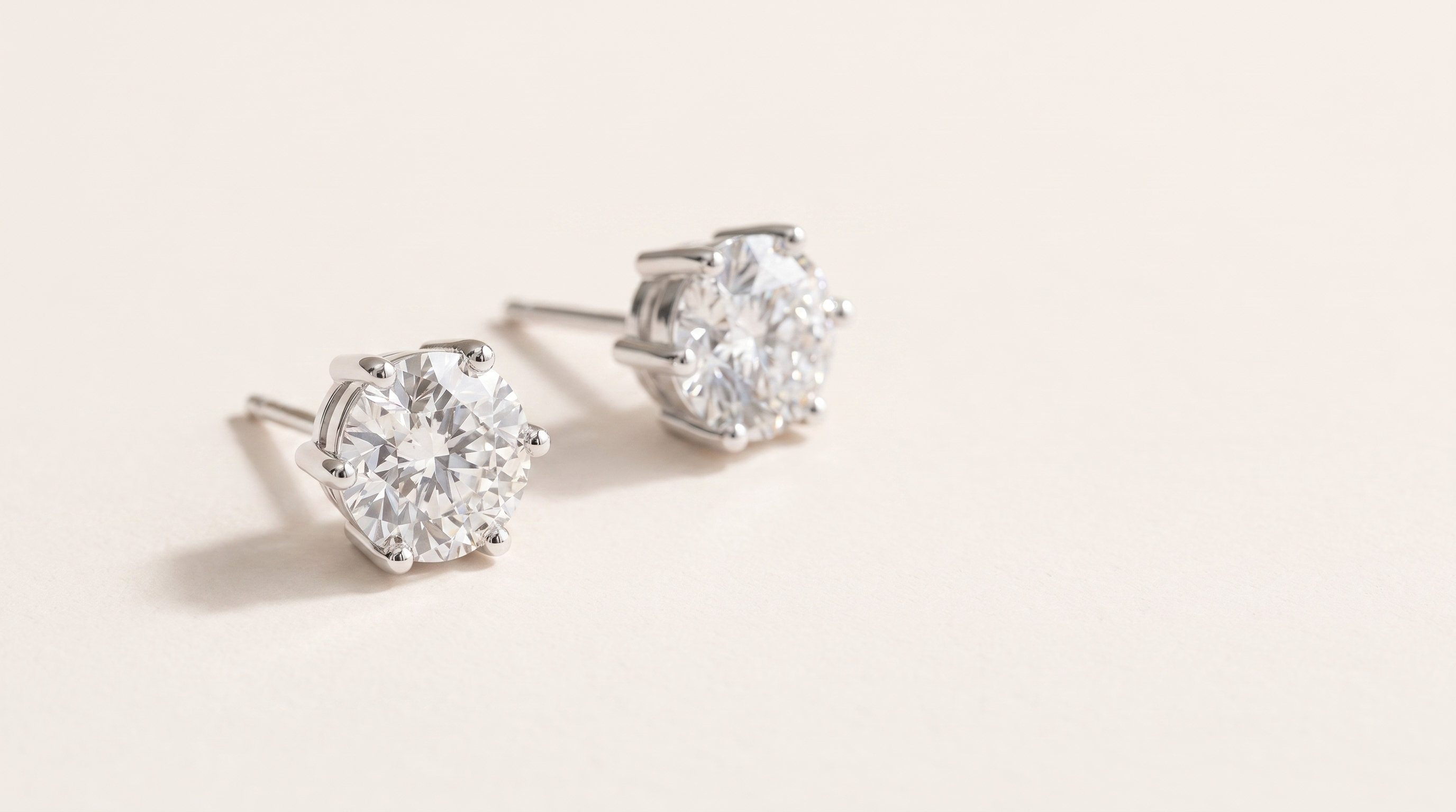 A pair of round moissanite stud earrings in six prong sterling silver settings, resting on a warm off white surface in soft daylight.