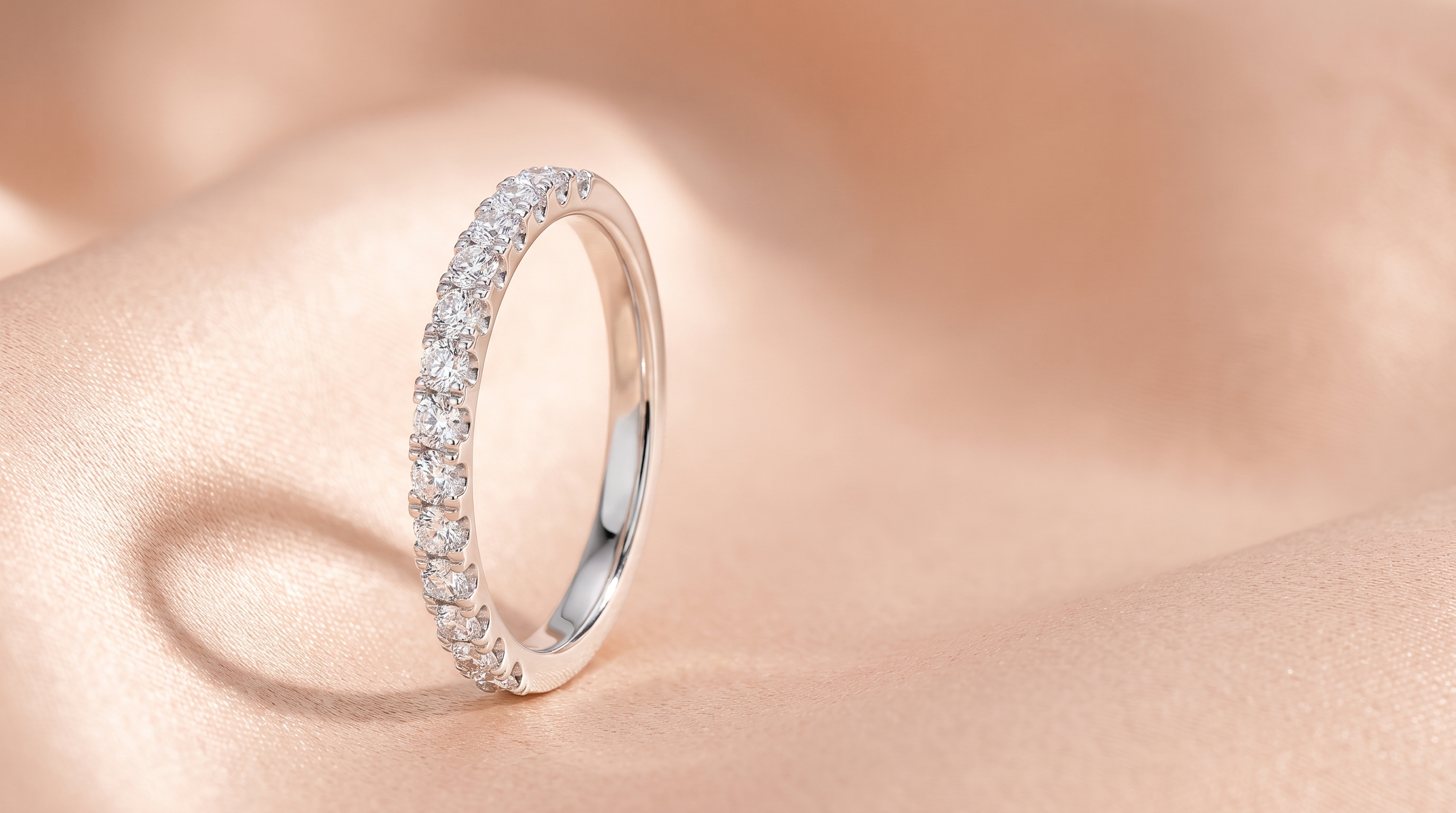 A slim silver half eternity band with a row of small round clear stones across its front, standing upright on folds of peach satin.