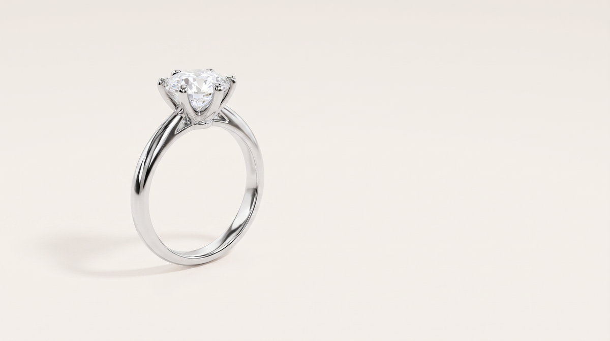 A silver six-prong solitaire ring with a round colourless stone, standing upright on a warm off white background.