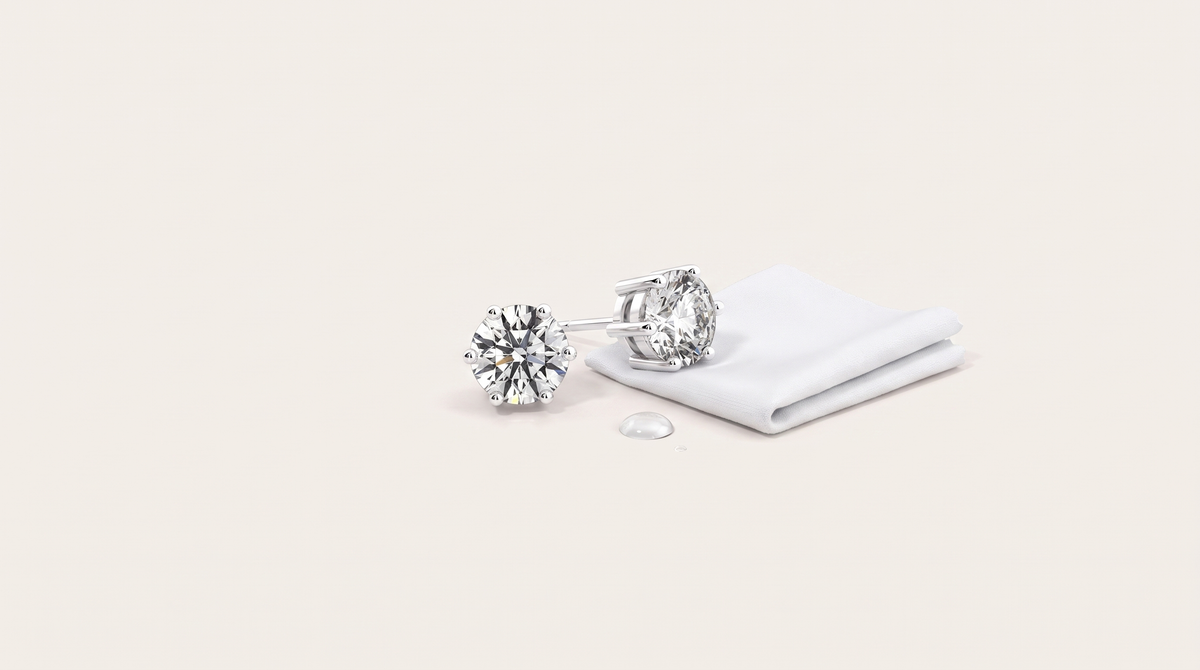 A pair of round six-prong moissanite stud earrings resting beside a folded white cloth and a single water droplet on a warm off white background.
