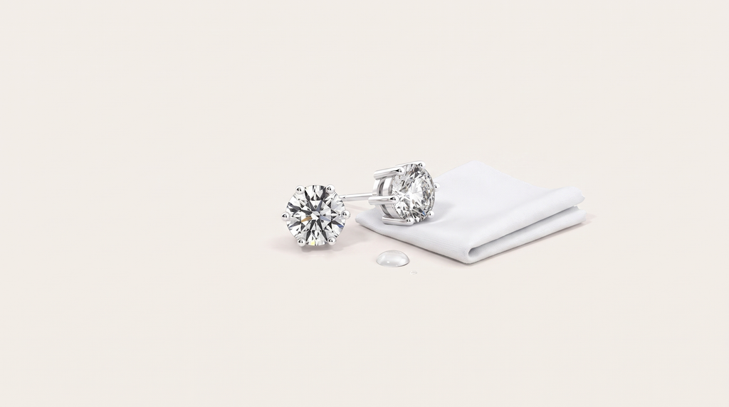 A pair of round six-prong moissanite stud earrings resting beside a folded white cloth and a single water droplet on a warm off white background.